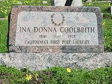A color photograph of a pink granite headstone amid lush green grass and yellow oxalis flowers. The headstone is engraved with a laurel wreath at the top, and reads "Ina Donna Coolbrith" 1841–1928 "California's First Poet Laureate".