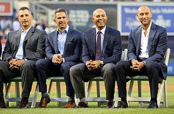 Andy Pettitte, Jorge Posada, Mariano Rivera, and Derek Jeter dressed in suits and seated in chairs on a baseball field.