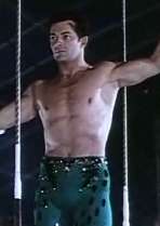 Frame from a film showing the torso of a bare-chested man standing on a circus trapeze; the man's arms are extended outwards from his body, and he's facing somewhat left of the camera.