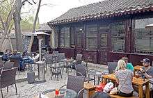 A courtyard surrounded by a high stone wall with a Chinese-style dark brown building at the rear. Within the courtyard are tables and chairs; people are seated and drinking beer at a few of them.