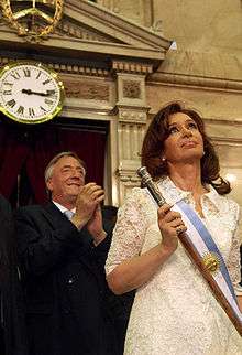 Cristina Fernández de Kirchner in formal presidential attire, including the presidential scepter. Husband and former president Néstor Kirchner stands behind her.