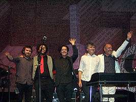 Five men are standing close together on a stage and smiling. First male at left is bearded and has right arm raised to shoulder height. Second male has arms around shoulders of his neighbours and is partly obscured by a microphone stand. Third male has left hand raised overhead. Fourth male has arms at side and is looking to his left. Fifth male has right arm over his neighbour and left arm overhead. Last two are partly obscured by a keyboard and its stand. Behind the five men is more band equipment and the background contains considerable English text.