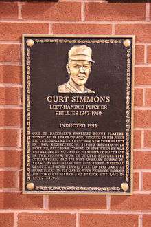 A bronze plaque mounted on a brick wall with the face of a smiling man wearing a baseball cap; the primary captions read "CURT SIMMONS; Left-handed pitcher; Phillies 1947–1960"