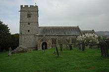 A square base of five steps made from large stones, supporting a base and a modern cross in front of a low stone church and tower