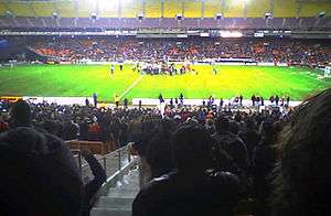 A team celebrates in the center of a soccer field while fans in stand on both sides cheer.