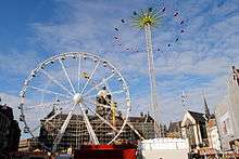 An image of the funfair in Dam square showing a Ferris wheel on the left, behind it spinning-arm ride called Speed; and a Matterhorn on the right with its spinning cars at maximum extension.
