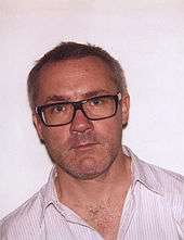 Damien Hirst, a man wearing glasses with brown rims, and a white shirt with faint vertical stripes
