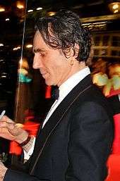 A man with messy black hair and a gold earring wearing a tuxedo and signing autographs.