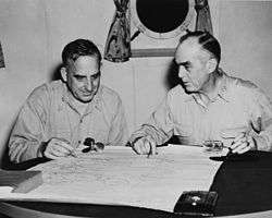 Two men sit at a small table. A map is laid out on the table. Its four corners are held down by a small propeller, a glass, and a book.