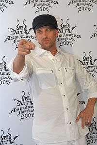 A white man in a white shirt with sleeves rolled up and a black baseball cap.  He is pointing at something in front of him with his right hand.