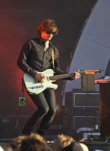 Middleton is playing his guitar with his left hand on the fret board and right hand on the strings. His knees are flexed, he wears dark glasses, dark pants and a dark shirt. His stage pass dangles from the end of the guitar. His hair is brown and hangs partly into his eyes. He wears a wide ring on his right hand's fourth finger. There is band equipment nearby.