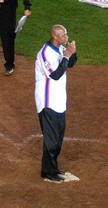 A man in black pants and a white baseball uniform with blue and orange stripes down the side holds his hands close to his face while standing on home plate.