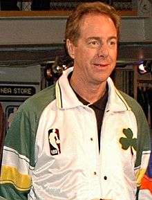 A man, wearing white and green jacket with the NBA logo and green shamrock on the front, is standing and looking to the front.