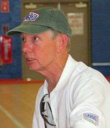 An older Caucasian man wearing a dirty green baseball cap and white polo shirt is sitting down on a chair in the middle of a basketball court. He is looking off to the left while conversing with a person off-camera.