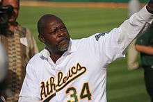 A bald black male baseball player in his fifties pitches a baseball. He is wearing an white uniform with the word "Athletics" across it, and the number 34 below the lettering.