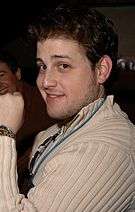 A dark haired Caucasian male wearing a beige colored sweater, with a blue colored lanyard and gold watch