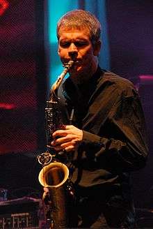 A man with his eyes closed, wearing a black dress shirt and playing a saxophone