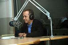 A man with headphones sits at a desk and leans towards a large microphone