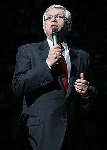 A man in his 50s with white hair speaks into a microphone. He wears a gray suit, red tie, and glasses.