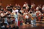 Five members of a band play keyboards, electric bass, drums and guitar in front of a symphony orchestra and its conductor, all dressed informally.
