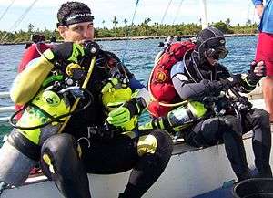 Two scuba divers in one-piece wetsuits and full scuba gear sitting in a boat, preparing to dive. Both divers are wearing tech rigs with back inflation BC and sling cylinders for decompression gas
