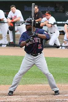 A man in a left-handed batting stance holding a black bat wearing a blue baseball jersey and grey pinstriped pants.