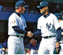 Rivera shakes Gary Denbo's hand wearing a white pinstriped baseball uniform with navy cap and undershirt.
