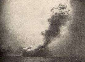 A black and white photograph showing a large cloud of smoke near the sea surface from which issues a towering mushroom cloud angled toward the right side of the photo