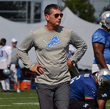 Candid photograph of Schwartz standing with arms akimbo on a football field wearing a grey long-sleeved t-shirt bearing a Detroit Lions logo, grey pants and sunglasses holding a whistle in his right hand and a small sheaf of papers in his left