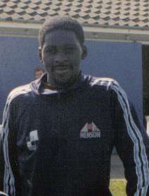A young man, wearing a black tracksuit top with blue and white stripes along the sleeves. Behind him is a building with a blue wall.