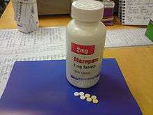 White bottle with red and black labels on a blue pad atop a desk. Also on the pad are seven small pills.