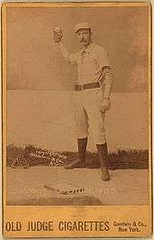 A baseball player is shown standing in his baseball uniform and gear used for a catcher.