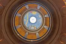The inside of a dome viewed from below. The first level's columns are topped with Ionic capitals, while we can see Doric capitals on the second level. At the center of the image is a mural depicting the New York state seal with planets, sun, stars and moon and a yellow ribbons across the sky. In the center is a circular skylight
