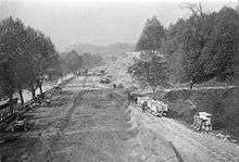 A wide dirt road beside a tree-lined river with several carriages and some vehicles