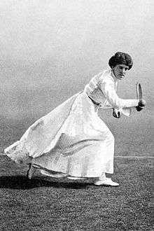 A black and white picture, a woman is in all-white attire hitting a right-handed one handed backhand with a racket in her right hand