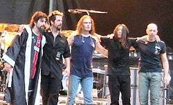 The five members of Dream Theater standing together in front of a drum kit and some amplifiers on a stage.
