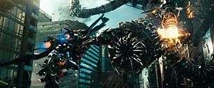 In a city full of skyscrapers, a robot wearing jet thrusters on his back holding a cannon flies toward a large snake-like robot. A tentacle of the larger robot is exploding.