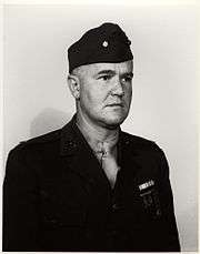 Head and shoulders of a white man wearing a garrison cap with an oak leaf on the side and a dark military jacket a row of ribbon bars and two badges on the left breast.