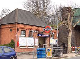 A red-bricked building with a blue sign reading "EAST ACTON STATION" in white letters and a man walking in front all under a white sky