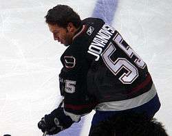 A Caucasian hockey player looking down at the ice while slightly crouched over. He is not wearing a helmet and is dressed in a black jersey with blue and maroon trim.