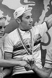 Eddy Merckx in a cap and white jersey with coloured stripes around the chest, holding his arm up