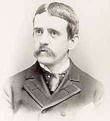A dark-haired, mustachioed man in his early forties wearing a black coat and tie