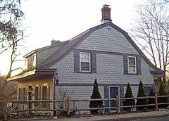 A gray wooden house seen from the side, lit by a setting sun from the left. Its roofline curves in segments, with a brick chimney at the crest. The upper section has wood shingled siding in a scale pattern.