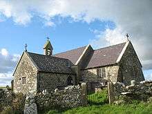A stone church with slate roofs seen from the southeast. To the right is the chancel, to the left is a large transept, beyond which can be seen a bellcote