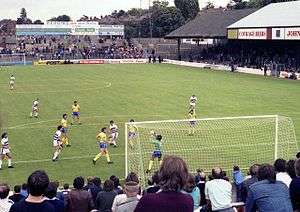 A colour shot of a football match in progress, taken from the terraced stand behind one of the goals. The terraced stand at the far end is without a roof, while the stand to the right is covered