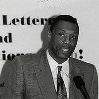 A picture of a darker black man wearing a suit while standing behind a podium. The photograph is in black and white.