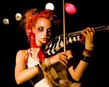 A red haired woman wearing heavy makeup plays a black and white striped violin.