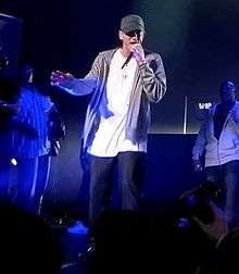 A stage picture of young man wearing a dark green cap, white shirt, brown jersey and dark pants singing into a microphone with neon blue stage lighting as his backdrop.