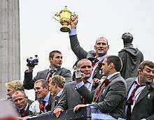 Photograph of nine members of the England rugby team on an open top bus victory parade. Lawrence Dallaglio is in the centre holding up the golden coloured Webb Ellis Cup, which is the trophy awarded to the winners.
