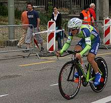 A cyclist riding a bicycle with a solid rear wheel and wearing a skin-tight lime green and blue jersey with white trim, with an aerodynamic helmet. Spectators watch him from the roadside.